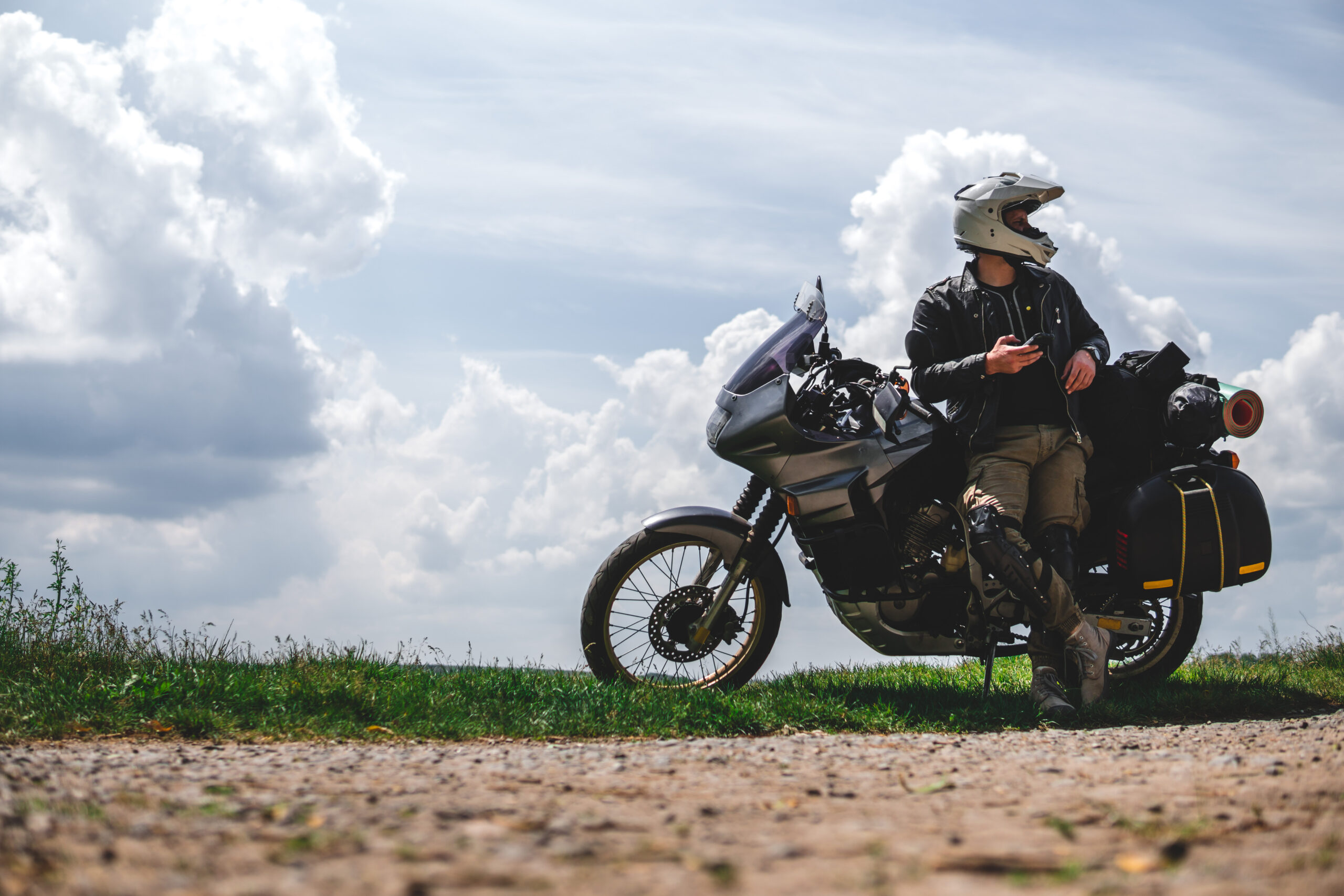En motorsyklist med hjelm og skinnjakke sitter på en fullastet motorsykkel under en himmel med dramatiske skyer, mens han ser til siden og holder en mobiltelefon i hånden.