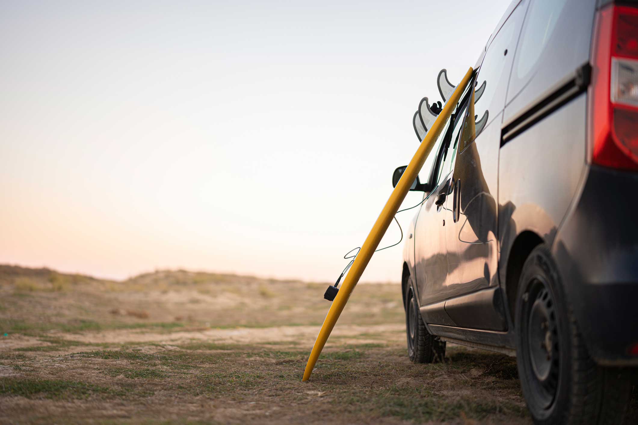 Et gult surfebrett står lent mot siden av en mørk varebil parkert på et tørt gressområde ved solnedgang.